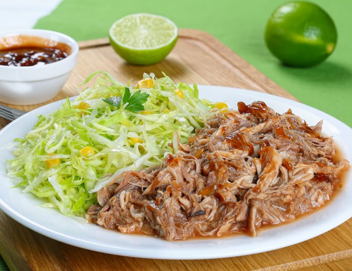 juicy pulled meat tossed in sauce on plate with fresh spring coleslaw corn salad served on wooden cutting board with fork and homemade barbecue sauce, view from above, close-up